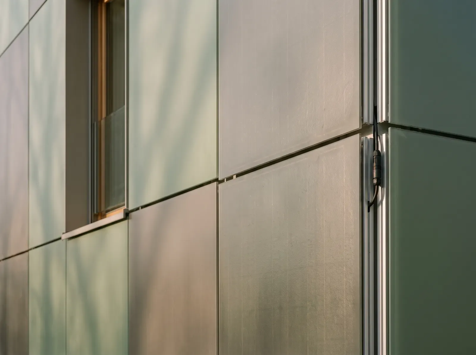 Nahaufnahme einer Solarfassade aus salbeigrünen und warmgrauen Glasmodulen mit satinierter Oberfläche, an der Kante die Aluminium-Unterkonstruktion und ein Steckverbinder