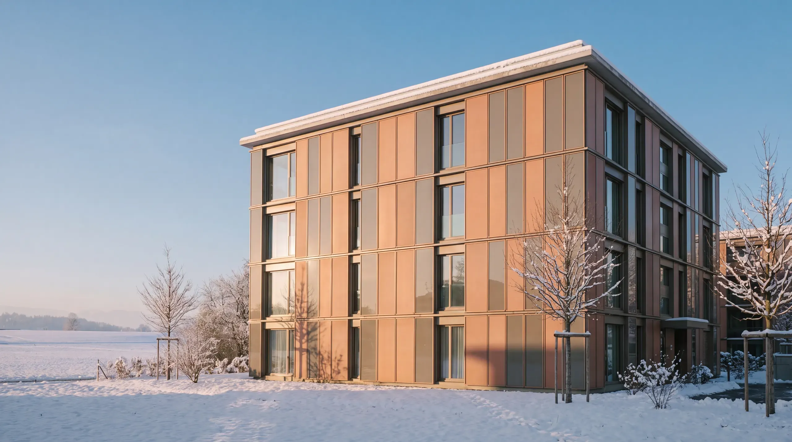 Vierstöckiges Mehrfamilienhaus im Winter, die Südfassade zwischen den Fenstern mit terrakottafarbenen und grauen Solarmodulen verkleidet, davor Schnee
