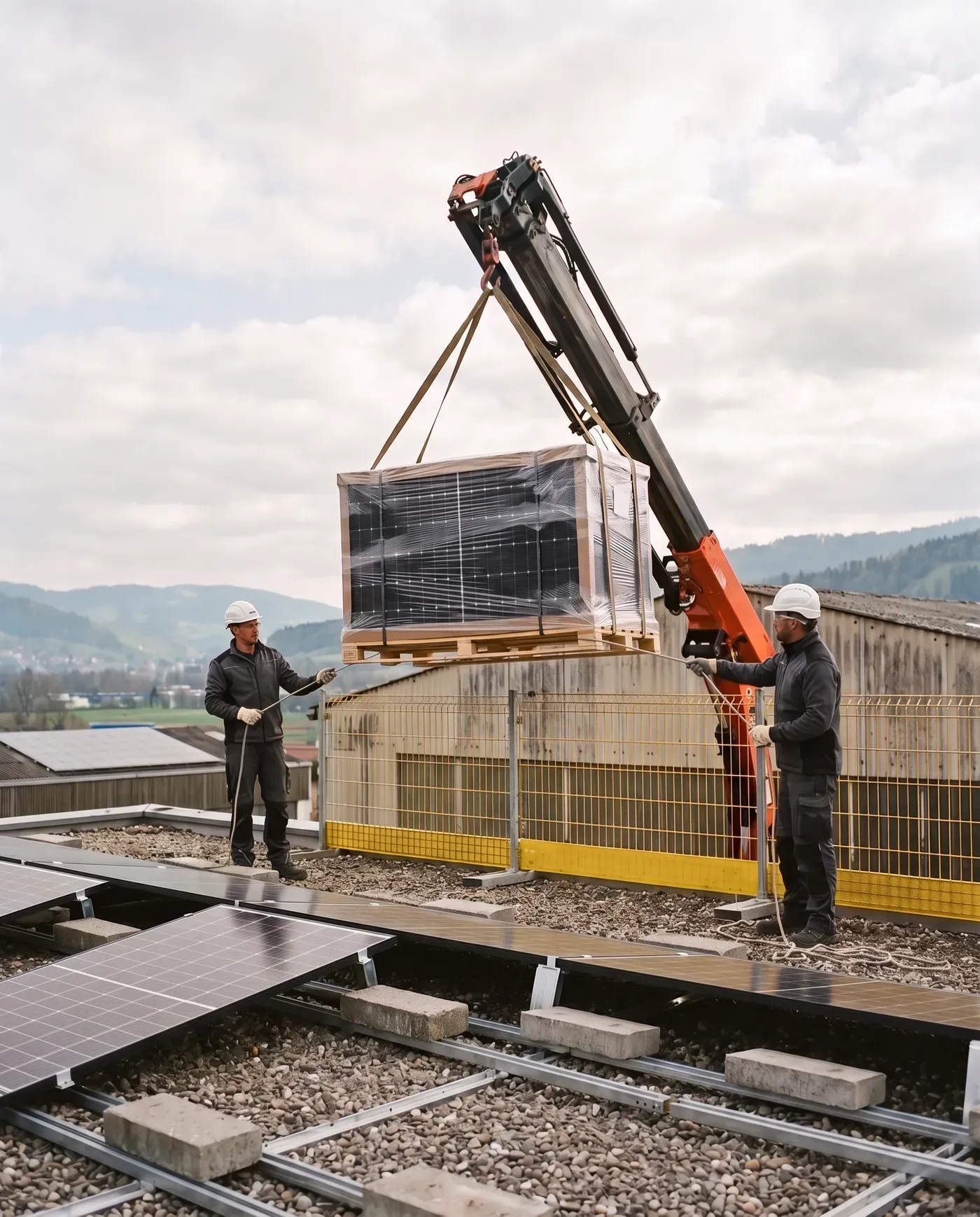 Ein Kran hebt eine Palette Solarmodule auf ein Flachdach, zwei Monteure mit Helm nehmen sie entgegen, am Dachrand ein gelber Seitenschutz